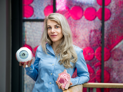 The Ph.d.-candidate in front of a pink glass painting, holding a medical eye model in one hand, and a mouth model in the other hand